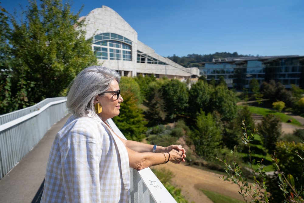 Businesswoman wearing glasses and a checked blazer stands on a balcony with metal railing, overlooking greenery and modern buildings under a clear sky, businesswomen, Spain.