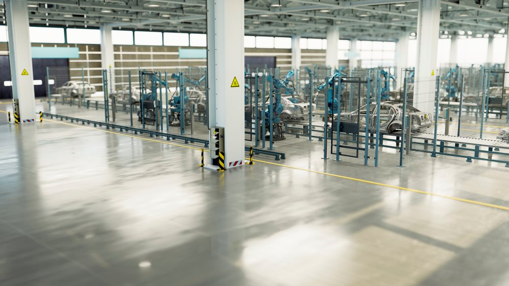 Car frames move along a conveyor under robotic arms inside a large industrial manufacturing factory.