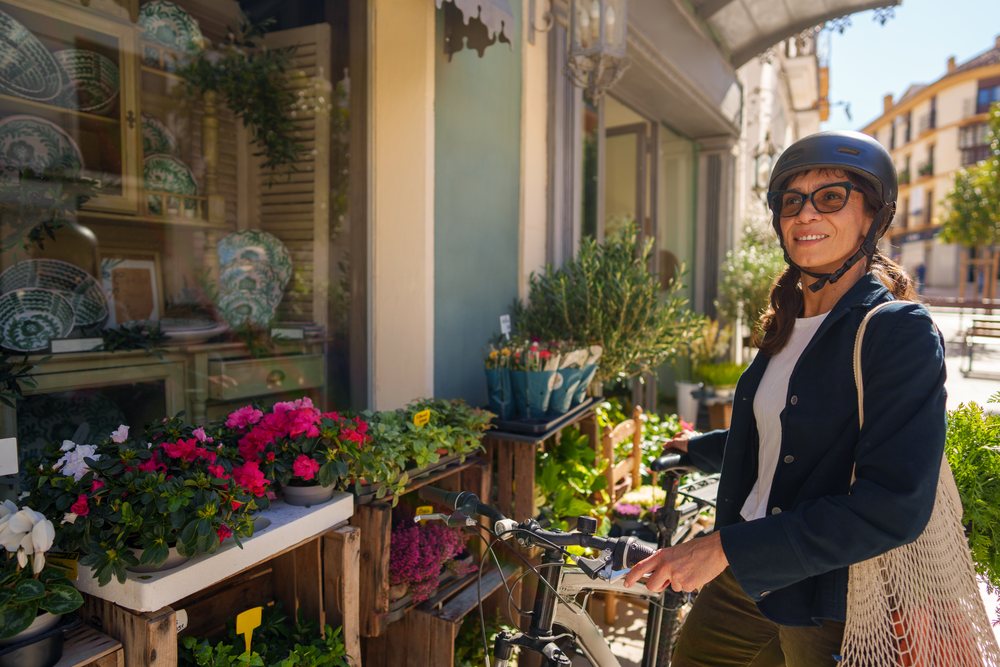 Woman wearing a helmet and eyeglasses stands holding a bicycle beside a flower shop with potted flowers on a sunny street, Málaga.