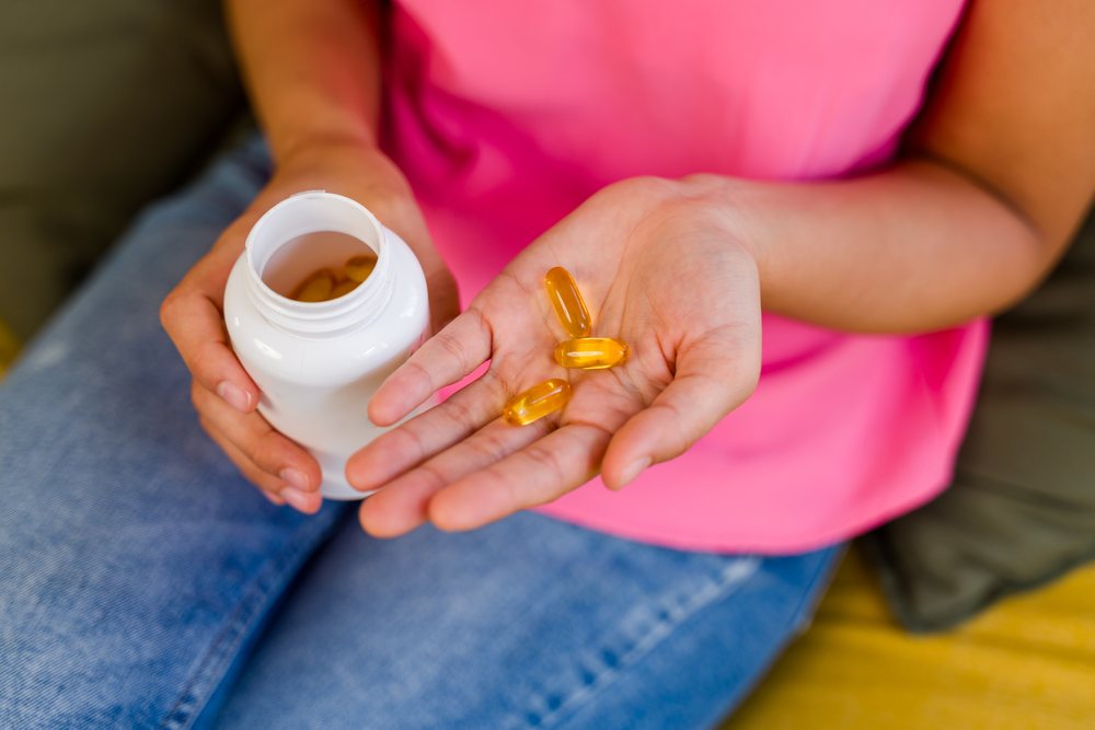 A person holding three yellow vitamin capsules in one hand and a white plastic container in the other hand in a casual setting, Spain.