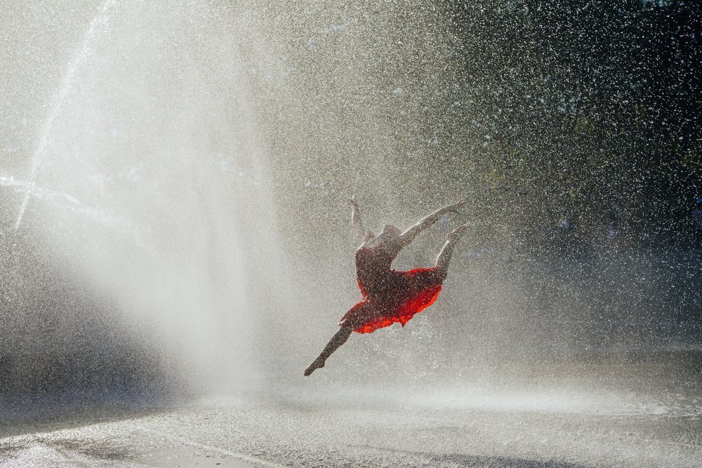 Female dancer in a red dress leaps dynamically through fountain spray outdoors, USA.