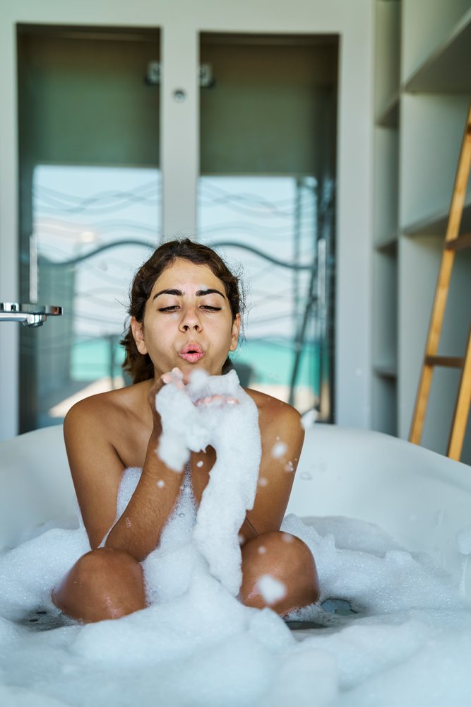 A young woman blows foam bubbles while relaxing in a bubble bath at home in the, Maldives.