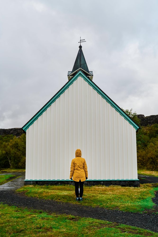 A person wearing a yellow jacket is standing facing the front facade of a small white church building with green trim and a steeple in a rural outdoor setting, Iceland.