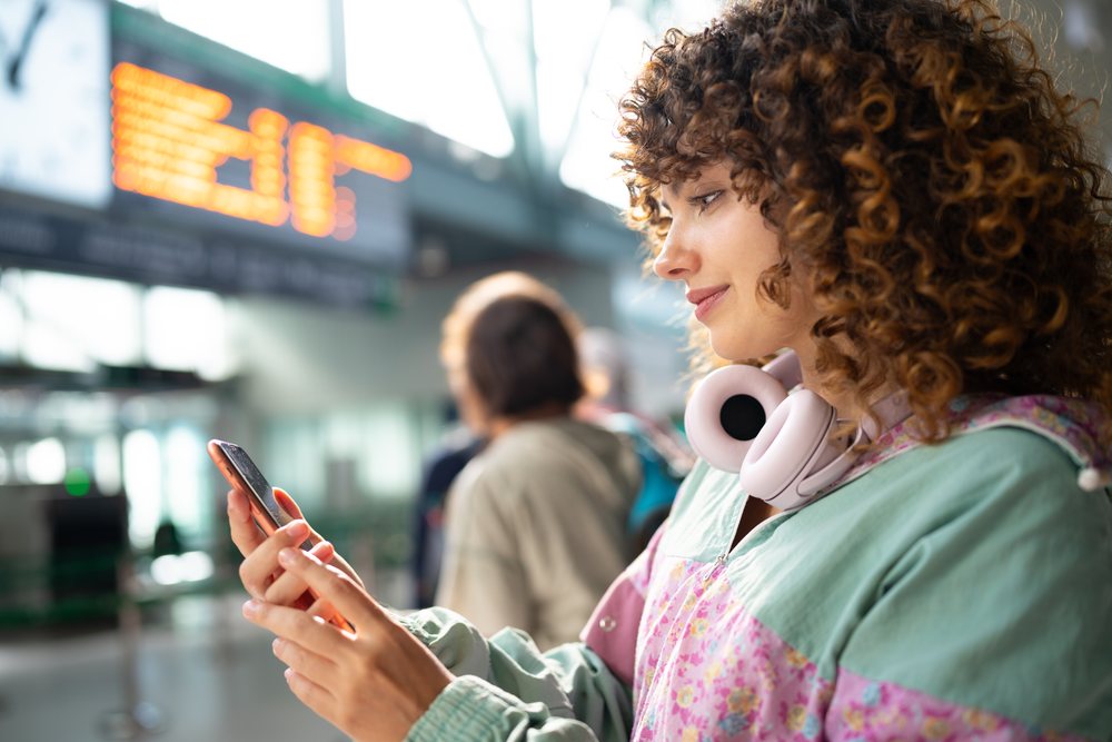 Woman with curly hair uses a smartphone at an indoor airport while wearing pink headphones around her neck, Spain.