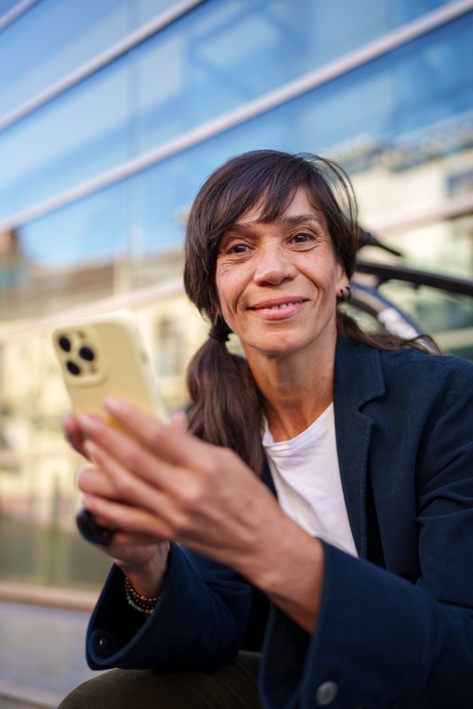 Woman wearing a blue jacket and white shirt sits outdoors near reflective building glass, holding a smartphone and smiling, Málaga.