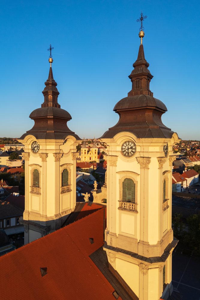 Two Minorite Church towers with clocks and crosses rise above red tile rooftops under a clear blue sky, Cityscape, Eger, Hungary.