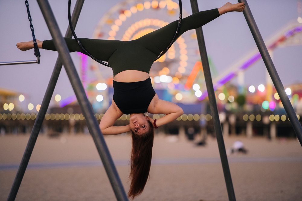 Woman hanging upside down on an aerial hoop outdoors at an amusement park during evening with a ferris wheel lit in the background, California, USA.