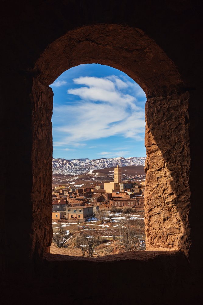 View through a stone window of Kasbah Glaoui showing the village of Telouet with snowy mountain terrain and a minaret tower, Lifestyle, Landscape, Culture, Morocco.
