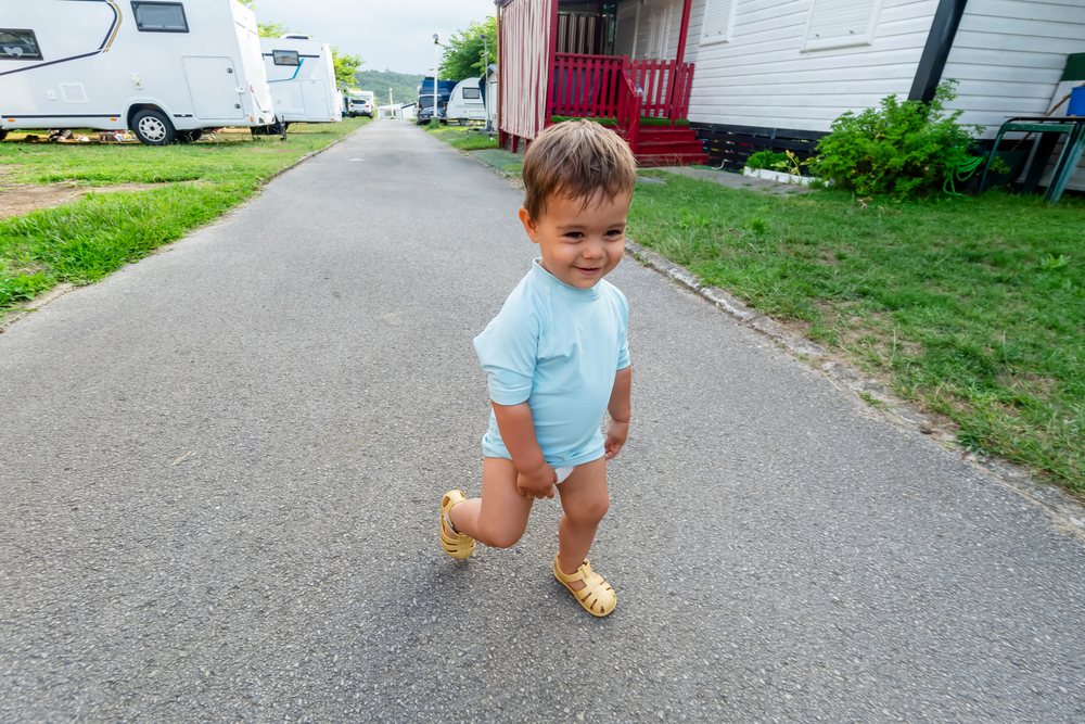 Toddler wearing a blue shirt and yellow casual footwear walks happily on a road beside camping trailers in an outdoor recreational area, kids, Spain.
