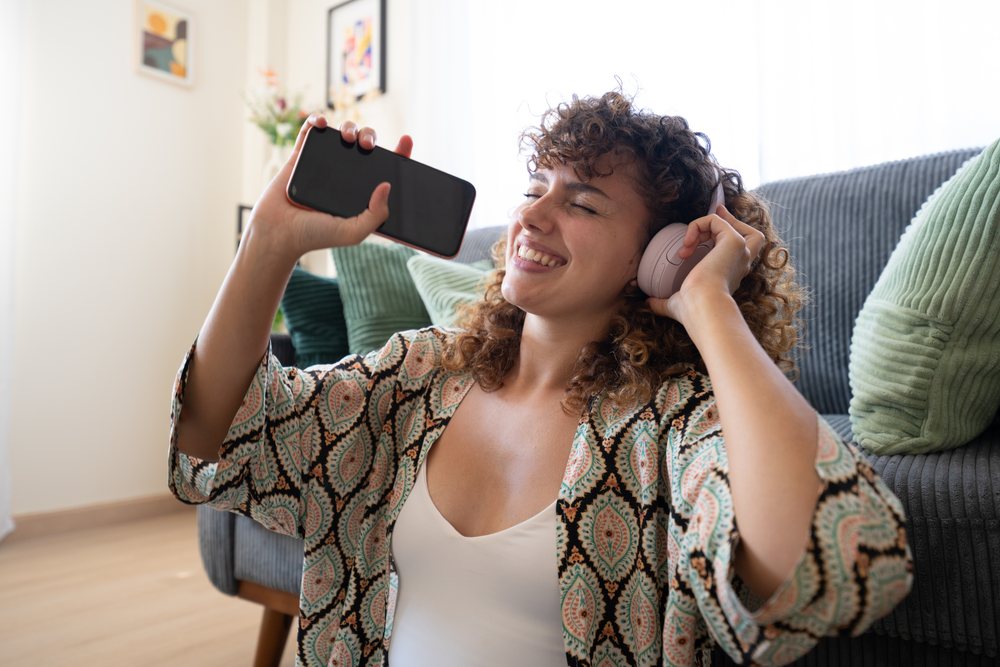 Woman with curly hair wearing headphones and holding a smartphone sits smiling on a couch indoors at home, Spain.