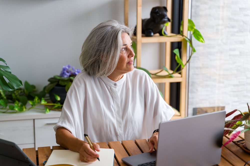 Mature gray-haired woman writing in a notebook while using a laptop at a wooden desk in a home office with plants visible indoors, Spain.