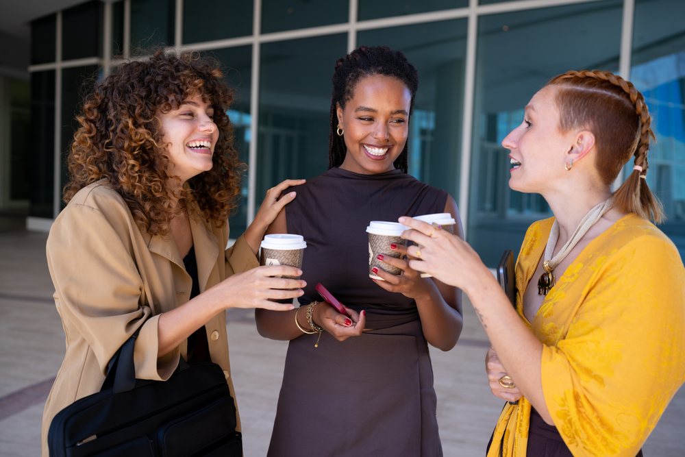 Three women holding disposable coffee cups laugh and chat in an urban outdoor plaza, one holding a smartphone, Spain.