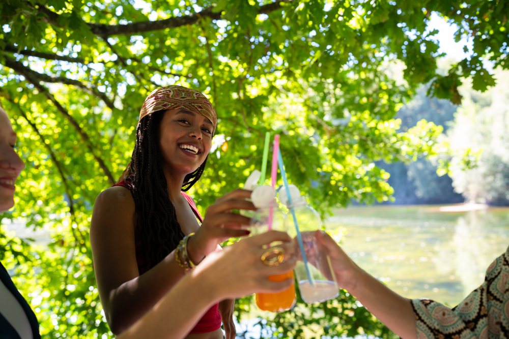 Friends celebrating outdoors with colorful drinks under tree shade near a lake on a sunny day, Spain.