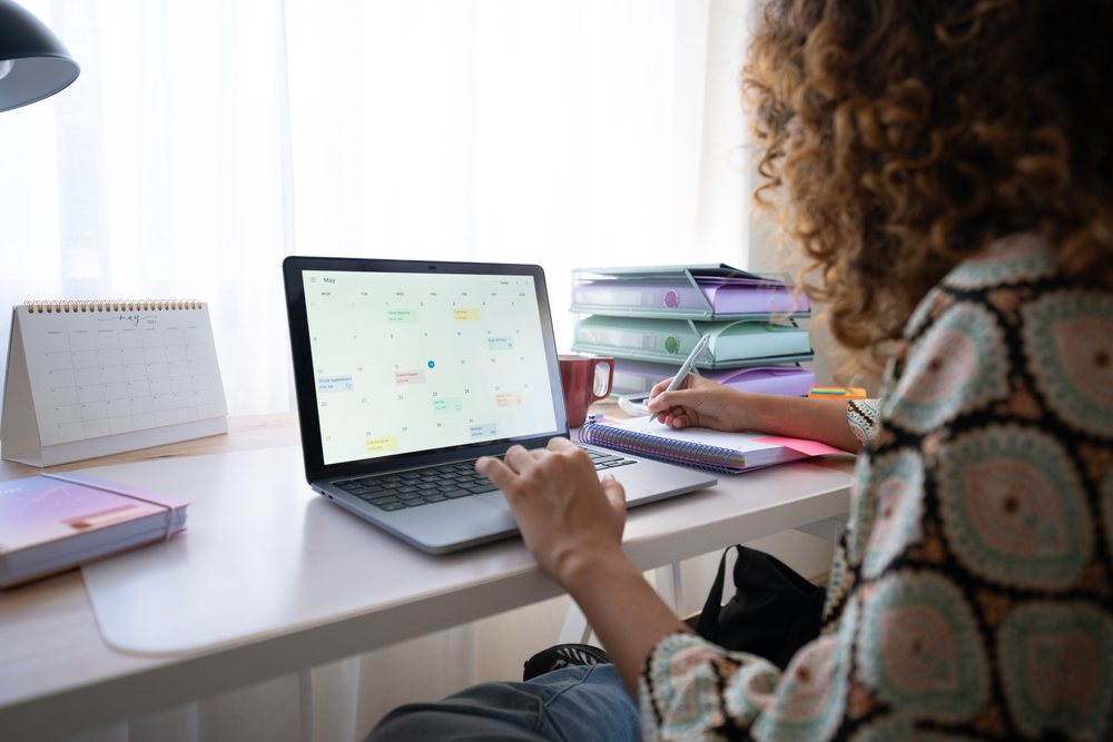Woman working at a home office desk with a laptop, calendar, and notebook, organizing her schedule in daylight, Spain.