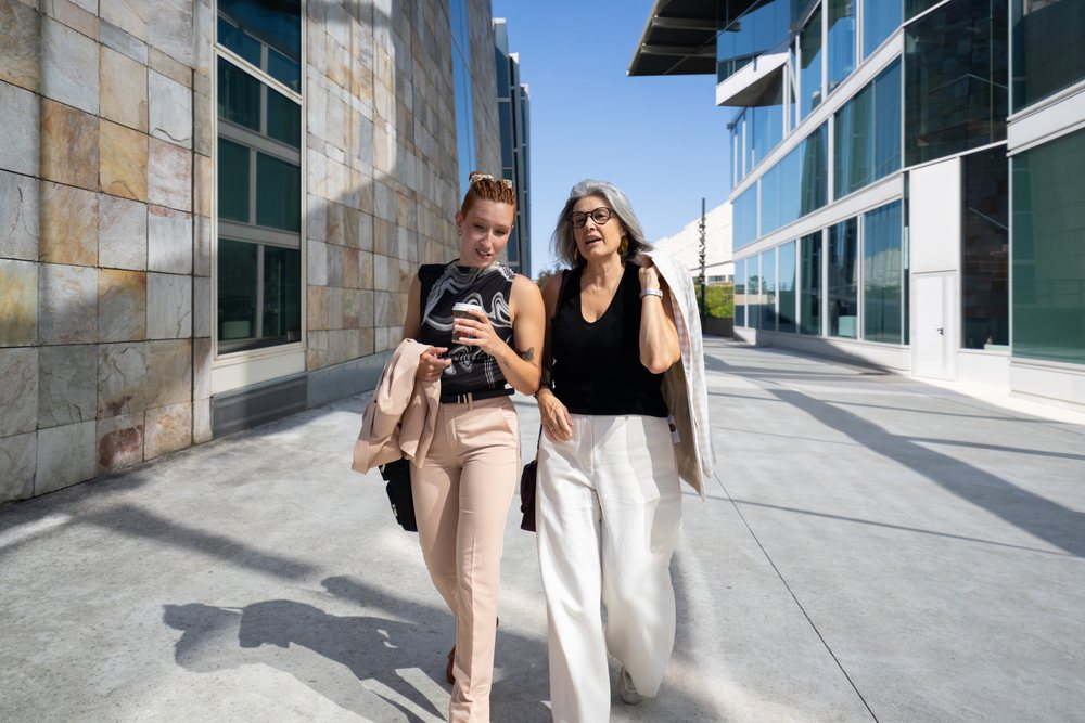 Two businesswomen walk and talk outdoors beside modern glass and stone buildings in sunny urban, Spain.