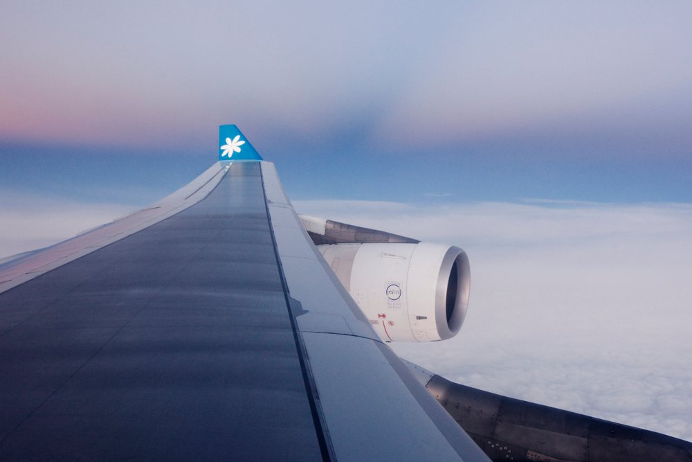 Airplane wing with engine and tail visible in flight above clouds under blue sky during sunset or sunrise, Tahiti.