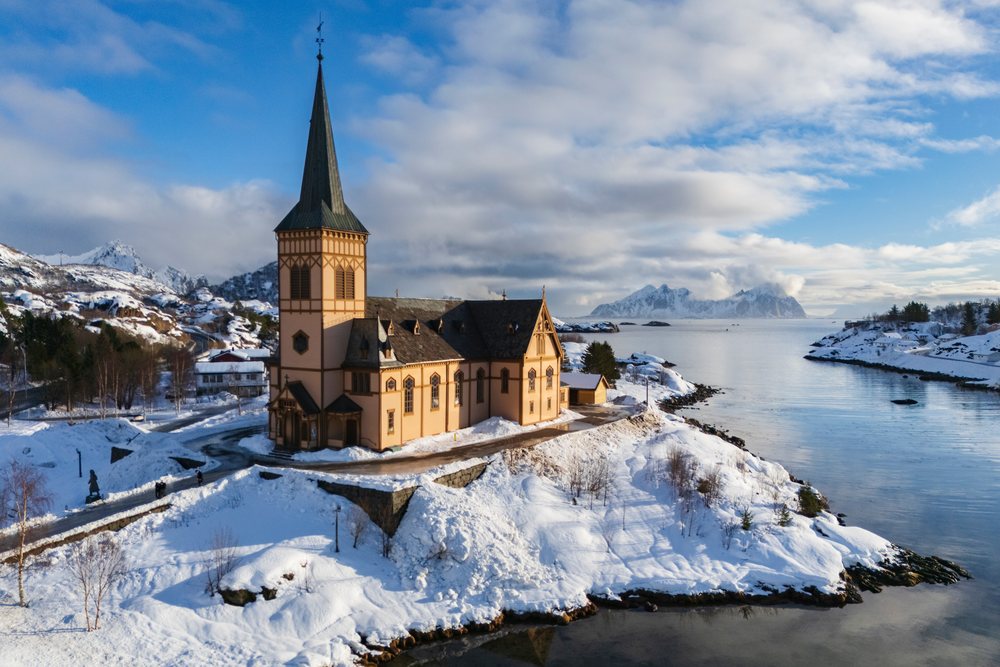 Vagan Kirche church with tall steeple beside calm water reflecting snowy mountains, Landscape, Kabelvaag, Norway.