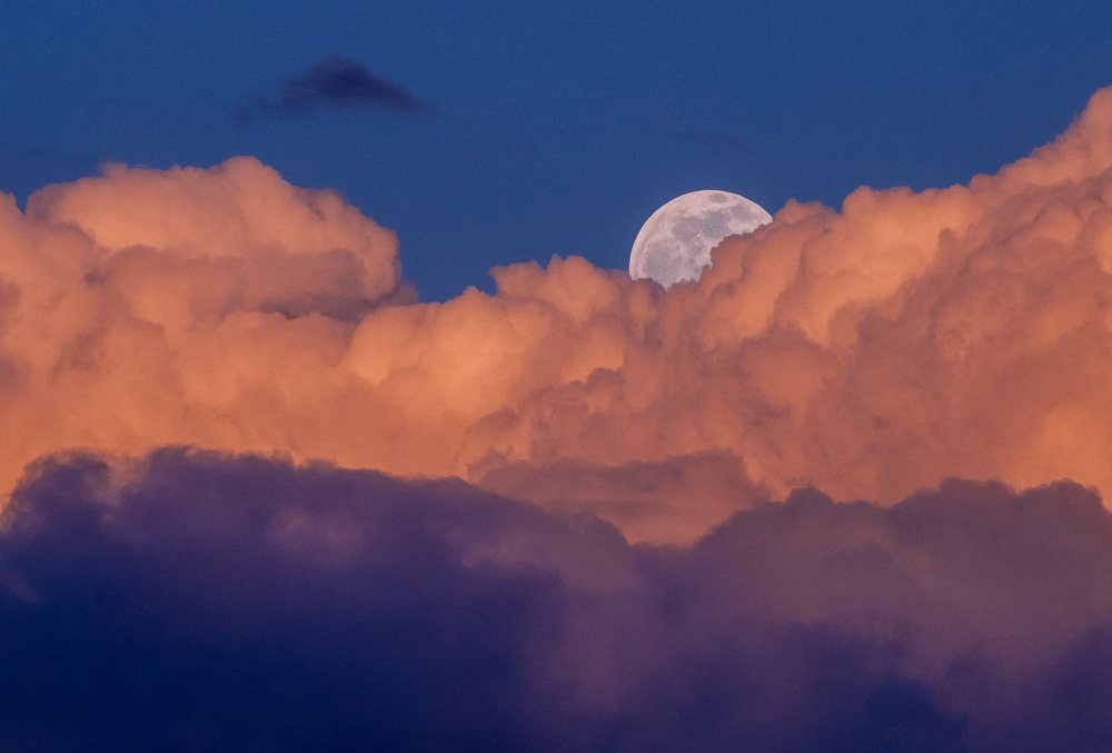 The full moon rises behind large orange cumulus clouds against a clear blue evening sky, Seattle, USA.
