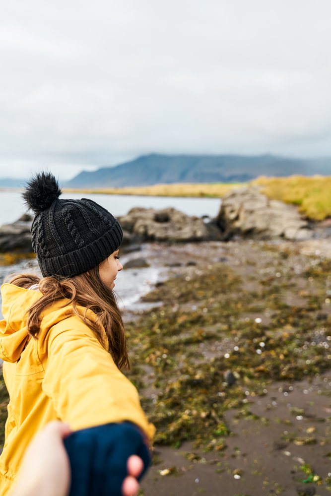 A white woman holding a hand walks along a rocky shoreline