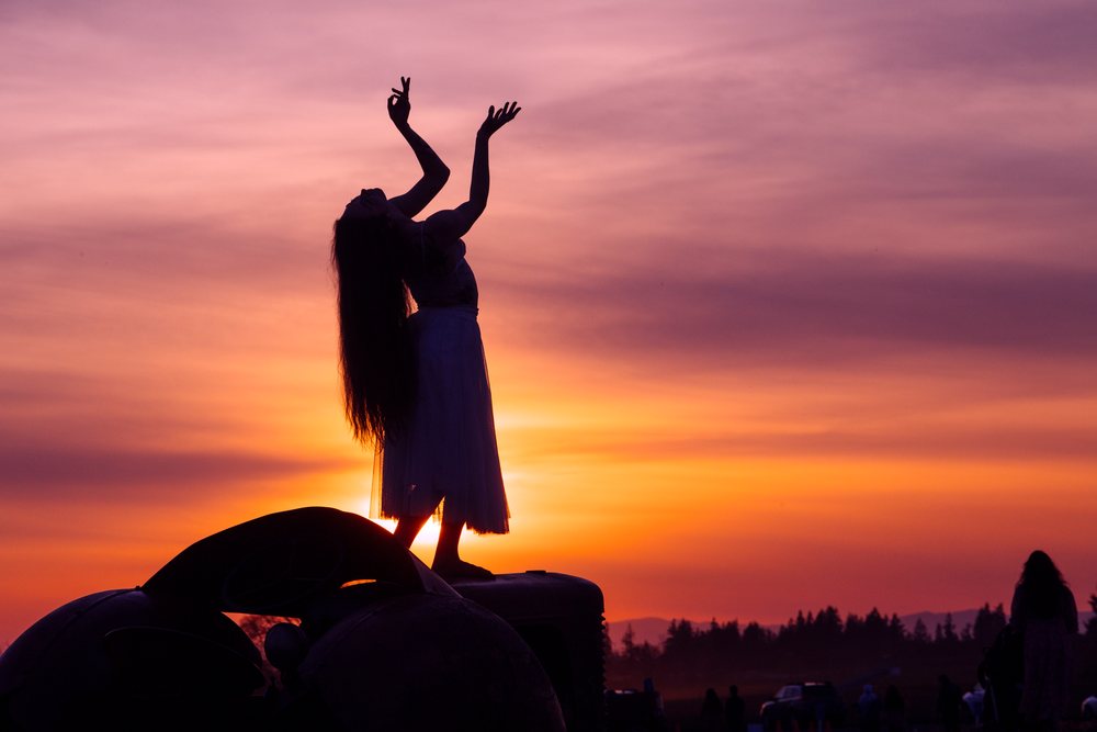 Silhouette of a female dancer with long hair and raised arms against a colorful sunset sky outdoors on a field, USA.