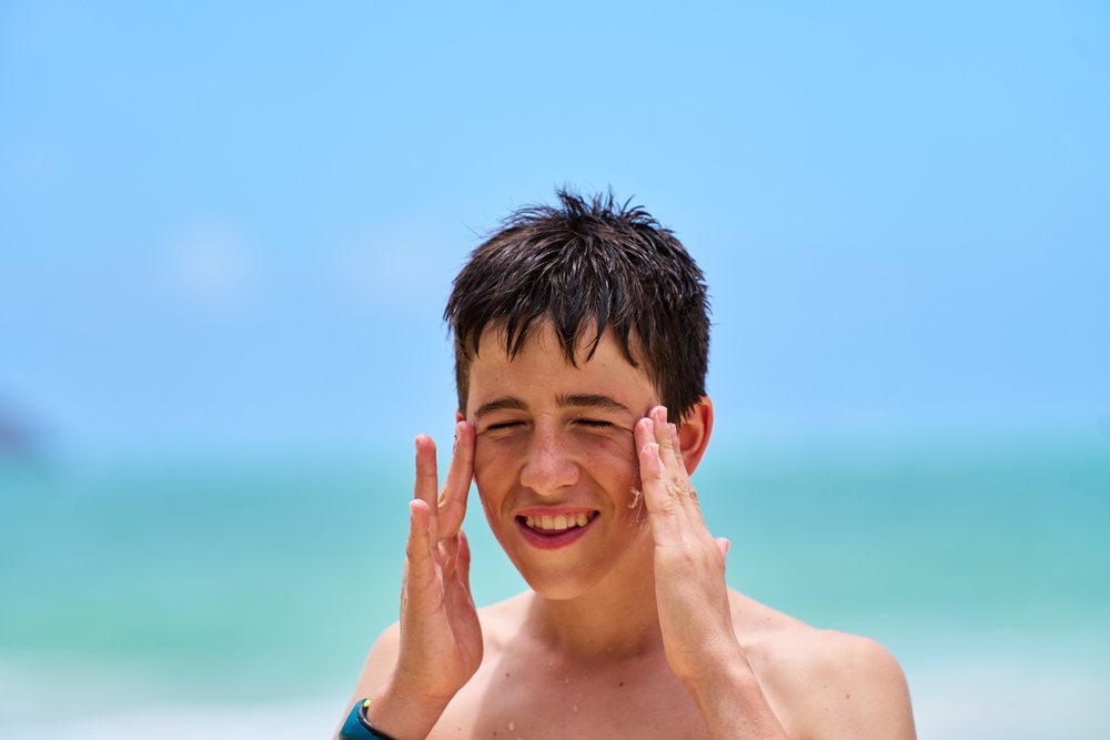 Boy applying sunscreen on his face at a sunny beach with ocean and blue sky visible, Hawaii, USA.