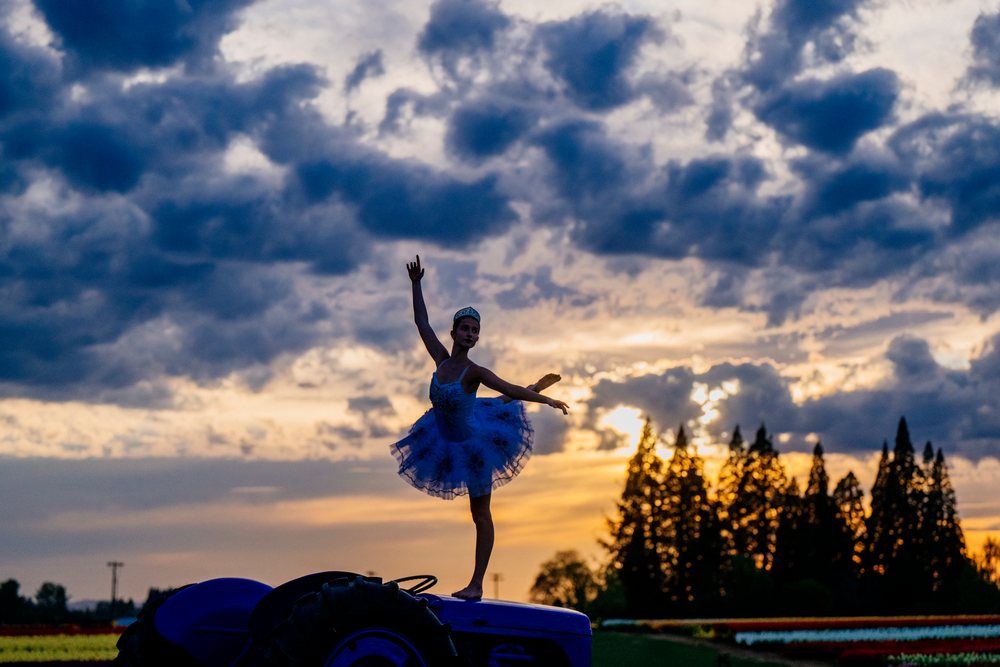 Ballet dancer wearing blue tutu and tiara poses gracefully on a tractor at sunset in a rural outdoor field under clouds, USA.