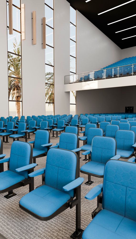 Empty modern auditorium featuring blue upholstered chairs and balcony seating, with large windows showing palm trees outside.