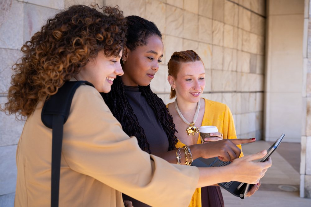 Three women smiling and collaborating outdoors near a stone wall while using a laptop and holding coffee, Spain.