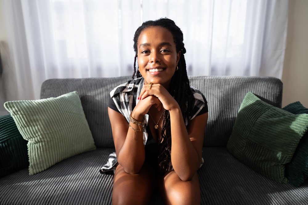 A smiling woman with dreadlocks sits relaxed on a gray sofa with green pillows in natural sunlight inside a living room, moving, Spain.