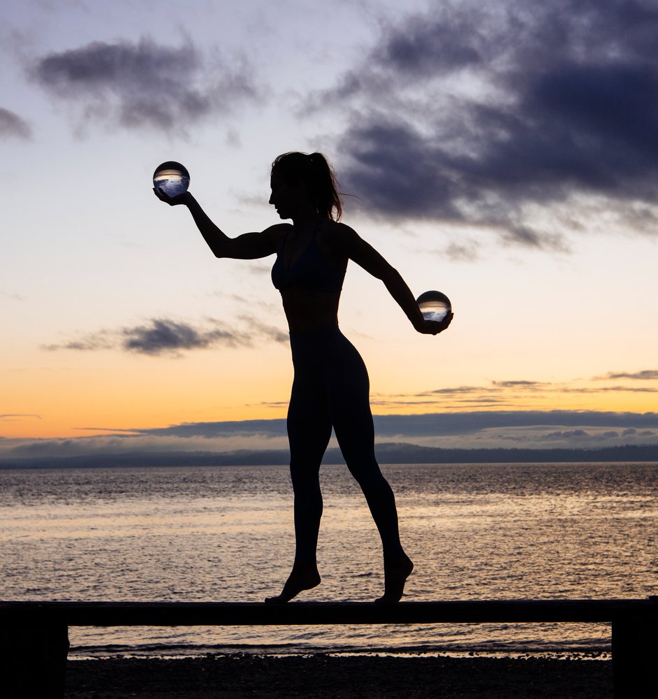 Silhouette of a woman balancing barefoot on a beam while holding two crystal balls on a beach at sunset, WA, USA.