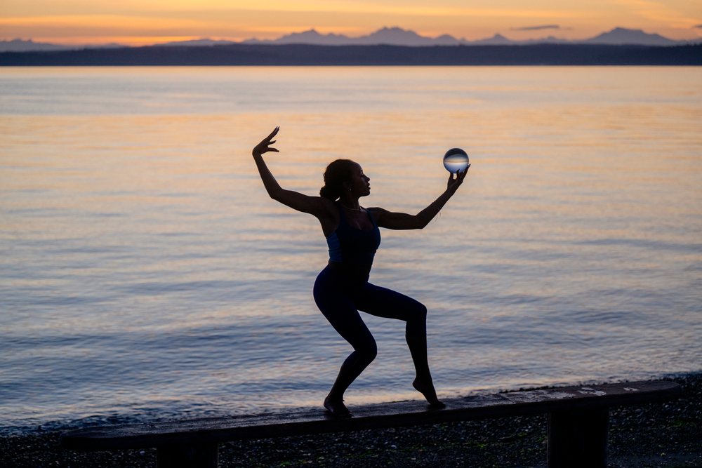 Woman practicing yoga acrobatics balancing on a bench near water at sunset, USA.