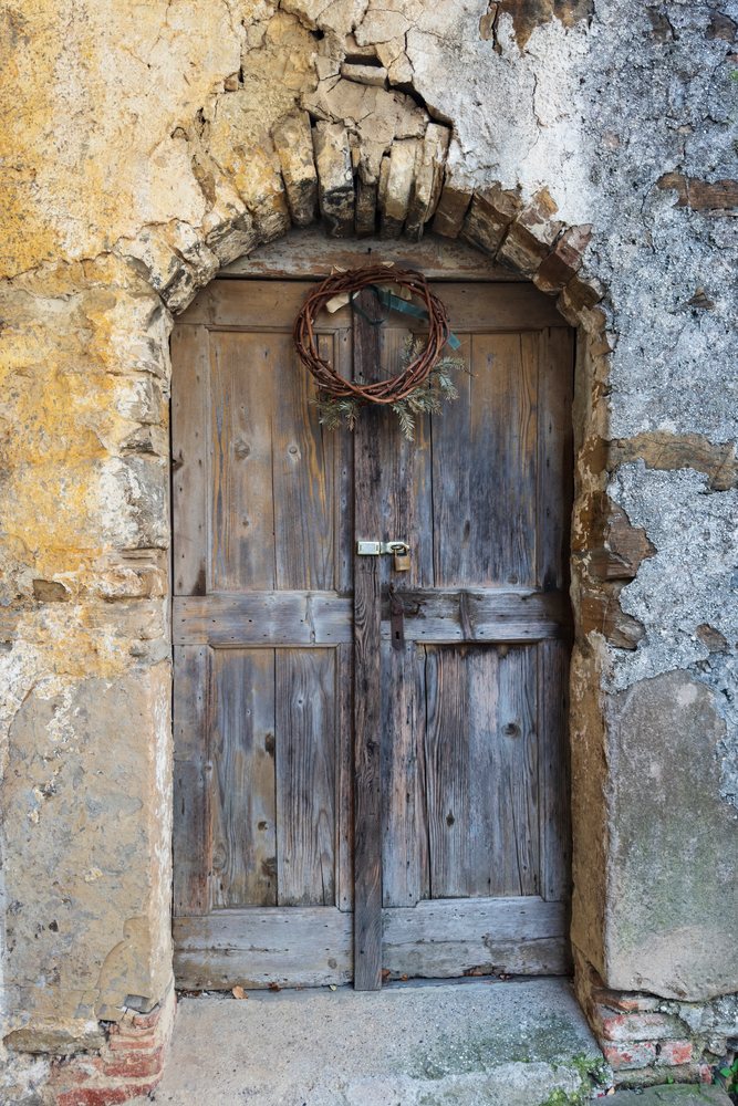 Rustic wooden door with a twig and pine leaf wreath hangs within an arched stone wall, secured by a lock and padlock, landscape, cityscape, Smartno, Slovenia.