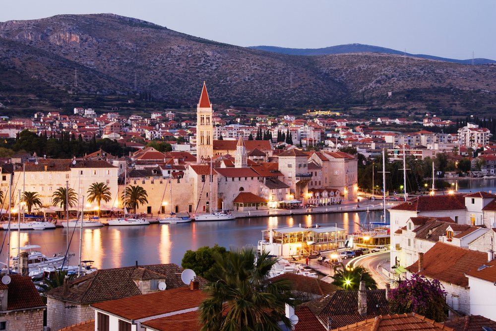 Town of Trogir featuring a harbor with boats and illuminated historic buildings under hills, Dusk, Croatia.