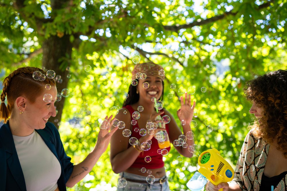Three women smiling and playing with bubbles outdoors under a leafy tree on a sunny day, Spain.