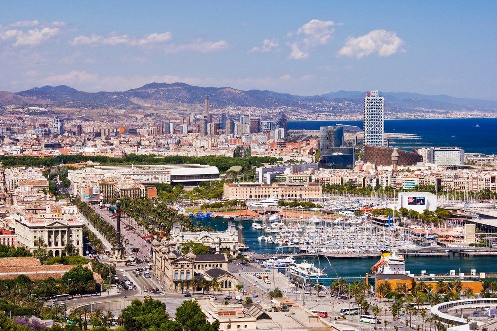 Barcelona cityscape showing harbor marina, urban skyline, and distant mountains under a blue sky.