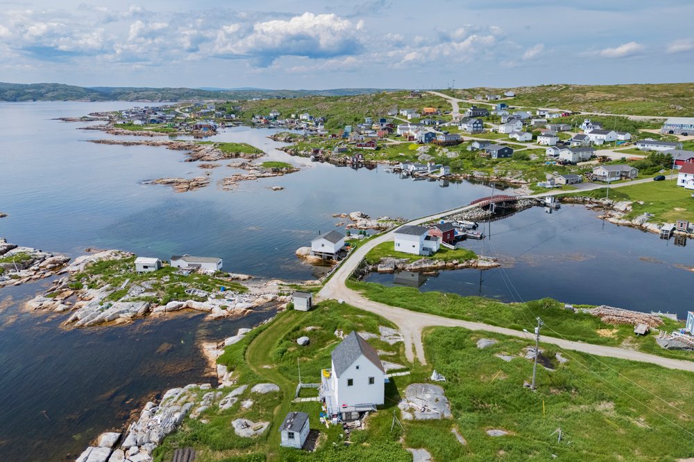 Aerial view of Greenspond coastal village showing small houses, a bridge over calm bay waters, rocky coastline, and green grassy areas under a partly cloudy sky, Landscape, Cityscape, Lifestyle, Newfoundland & Labrador, Canada.