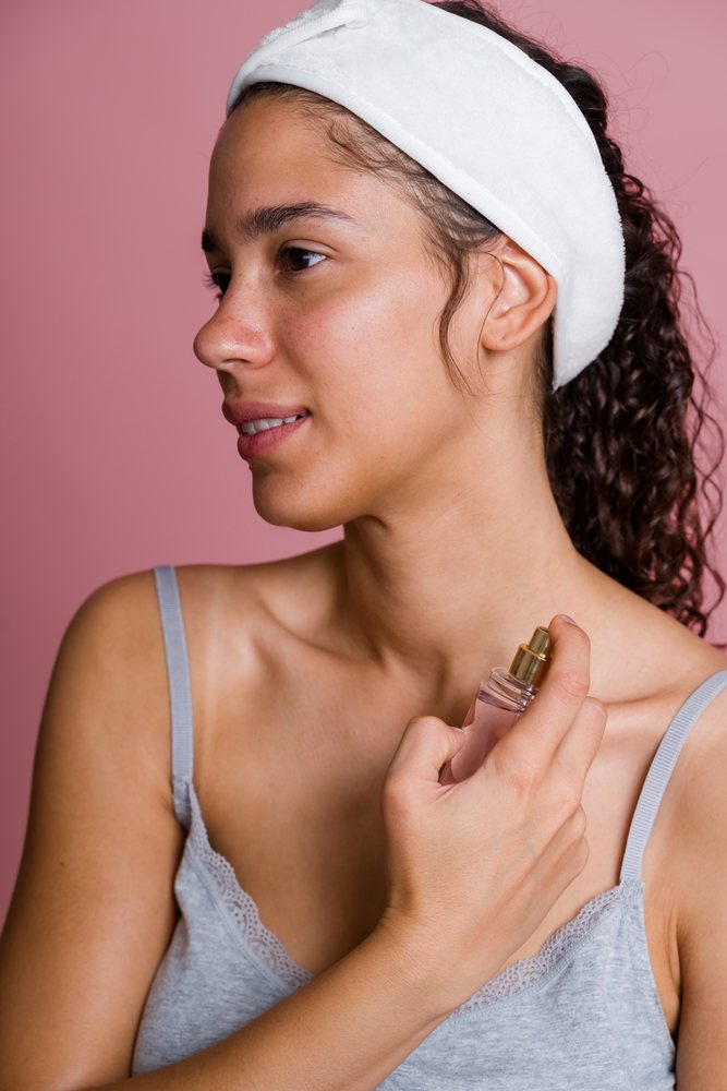 Woman with curly hair wearing a white headband sprays facial mist on her natural, skin in a pink beauty shoot studio, Spain.