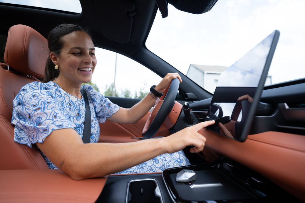 Smiling woman in a blue patterned dress uses the touchscreen dashboard while seated and belted in the driver's seat of a modern car with brown leather seats, Spain.