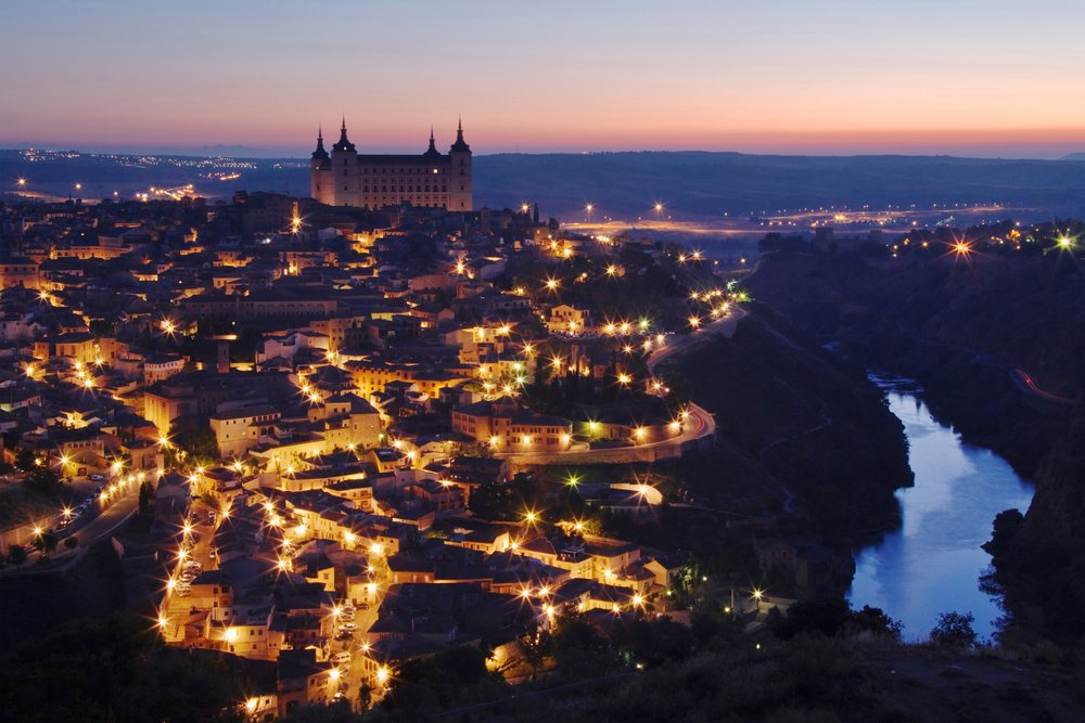 Evening view of Toledo cityscape with illuminated castle and river flowing beside the city.