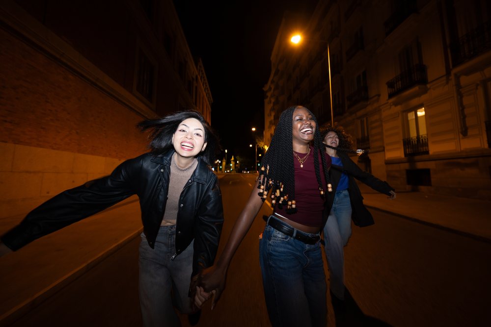 Three women holding hands and laughing as they run along a city street at night, Madrid, Spain.