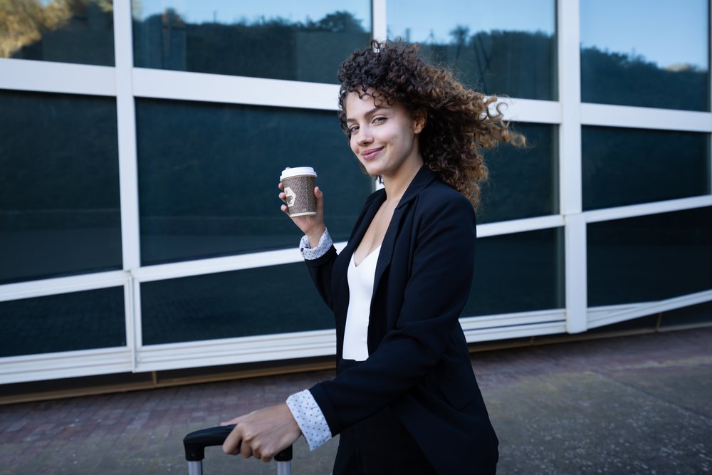 Businesswoman holding a coffee cup and pulling a suitcase while walking outside a modern building during a business trip, Spain, Santiago De Compostela.