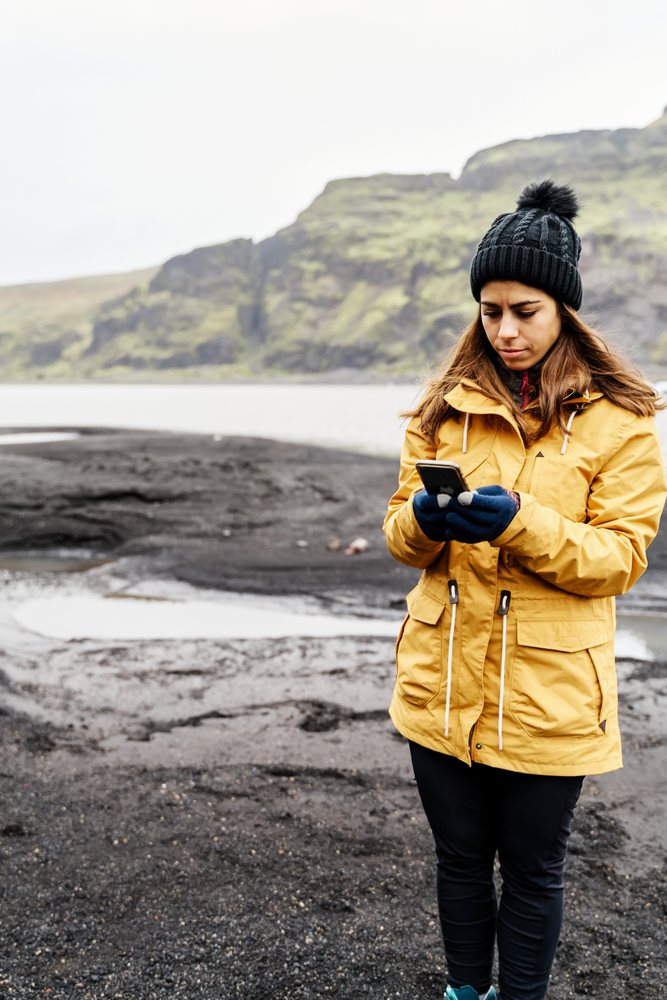 A woman wearing a yellow jacket and black beanie uses her smartphone while standing on rocky terrain with a mountain landscape, Iceland.