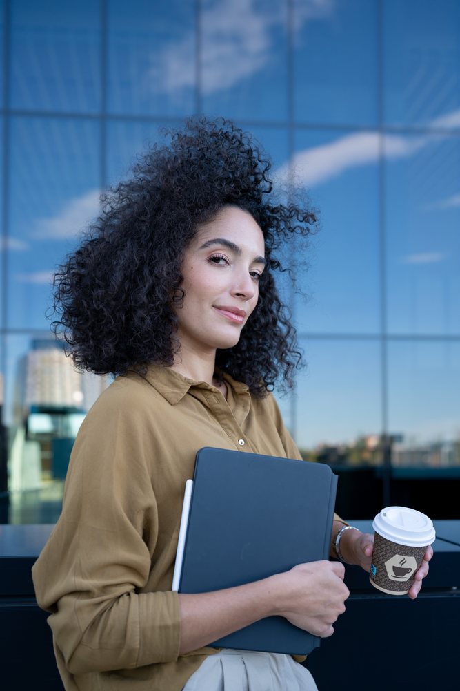 Woman with curly hair wearing a brown shirt holds a tablet and coffee cup outside a glass office building, Madrid, Spain.