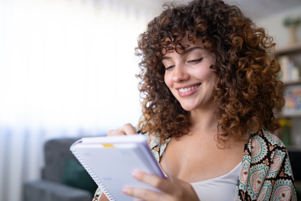 Woman with curly hair is smiling while writing in a spiral notebook in a casual indoor home setting, Spain.