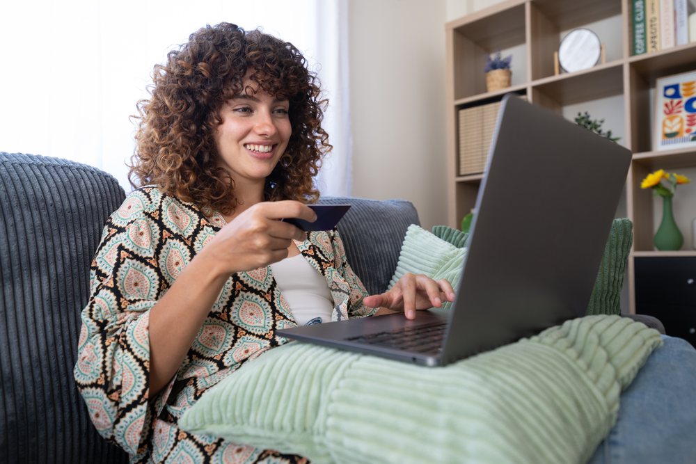 Woman with curly hair shopping online using a laptop and credit card while seated on a couch in a daylight-filled living room, home, girl, Spain.