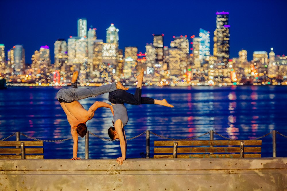 Two people perform partner acrobatics handstands by the Seattle waterfront at night with illuminated skyline and reflections in water, USA.