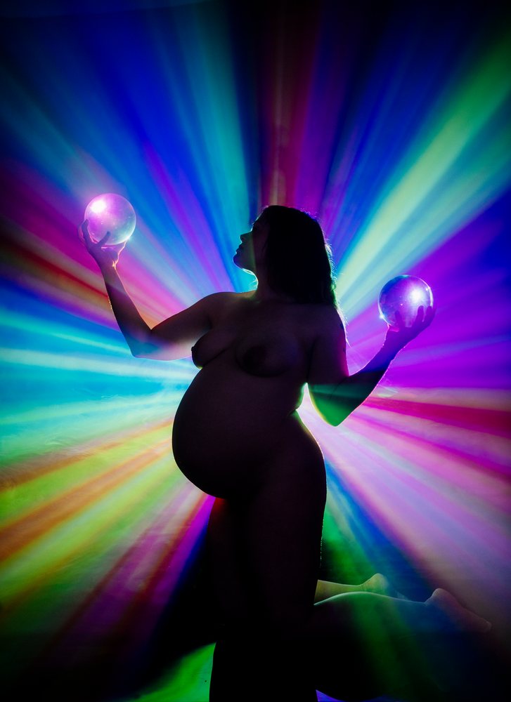 Pregnant woman kneeling in silhouette, holding glowing spheres with vibrant rainbow rays in a dark background, pregnancy.