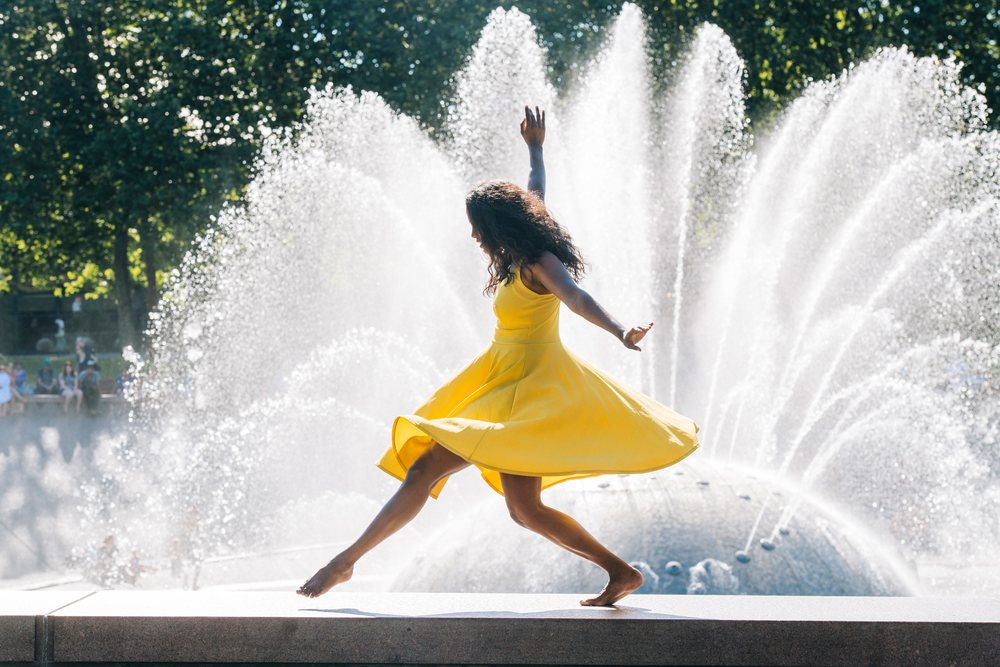 Woman dancing barefoot in a yellow dress by a large water fountain outdoors in a park with grass and trees under sunlight, USA.