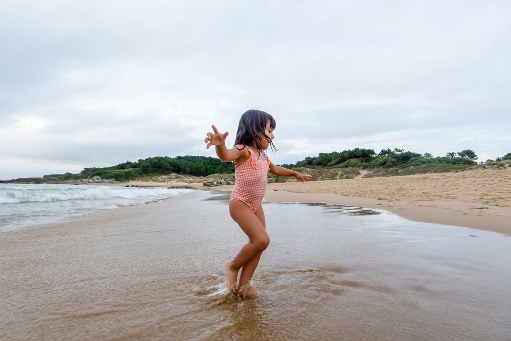 Child girl in pink swimsuit playing in shallow water on sandy beach coast in Spain under cloudy sky, kids.