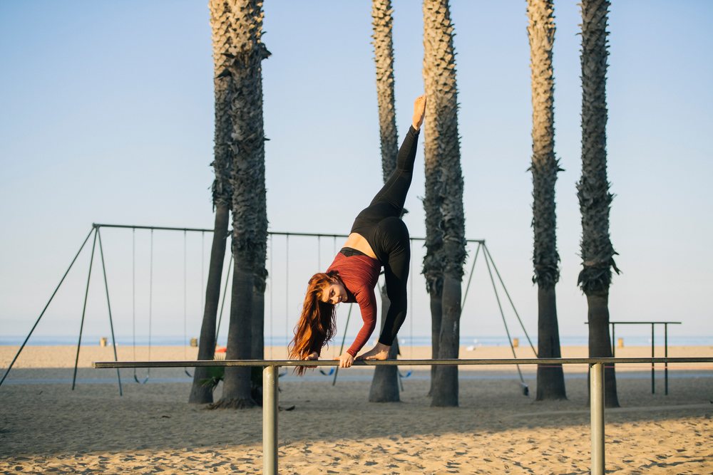 Woman performing balancing exercise on balance beam at sandy beach with palm trees and swings, California, USA.